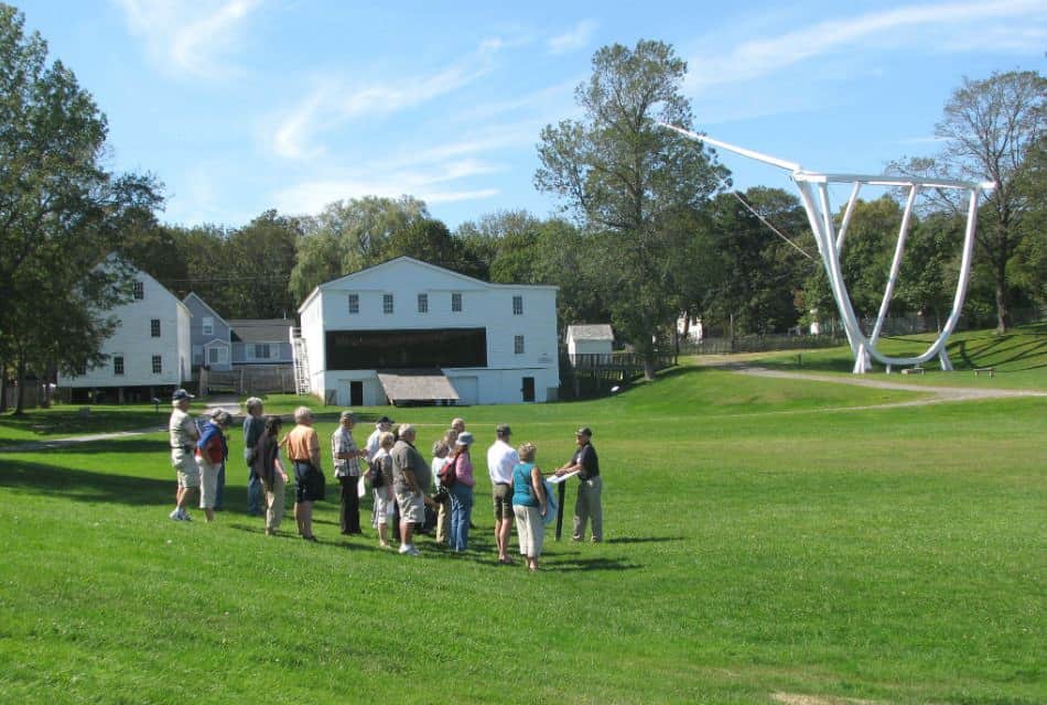 A man standing at a podium talks to a group of people on a green lawn next to large white structures.