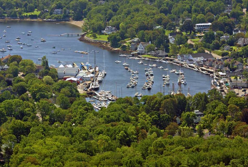 Many boats fill a harbor surrounded by lush green trees and homes next to a sandy beach.