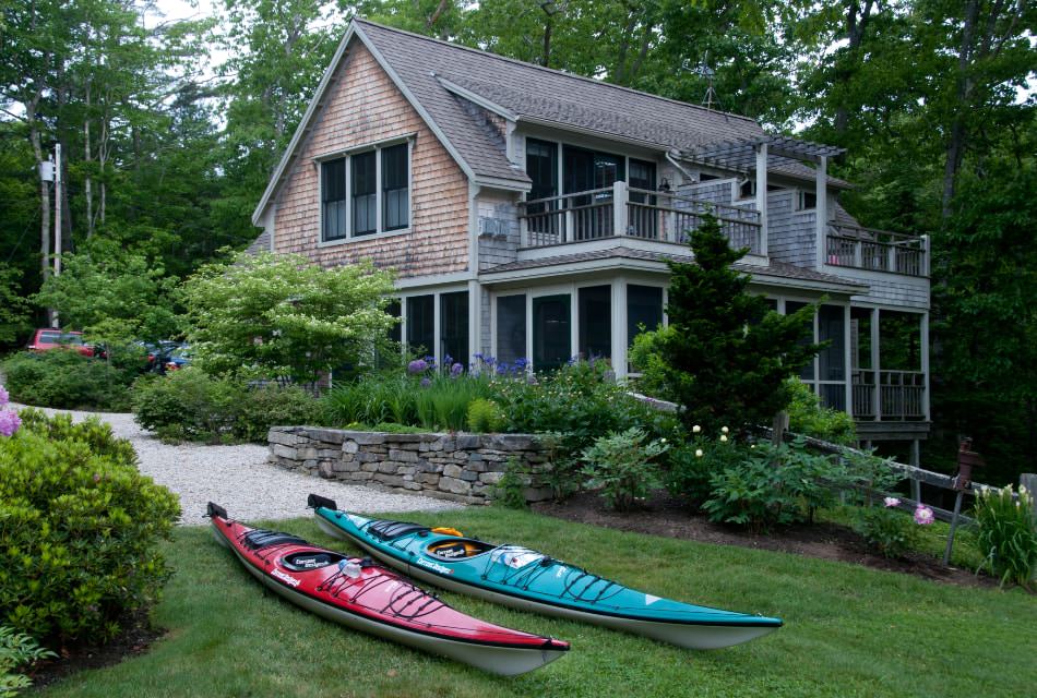 A red and a teal kayak lie on the grass outside a two-story house with balconies.