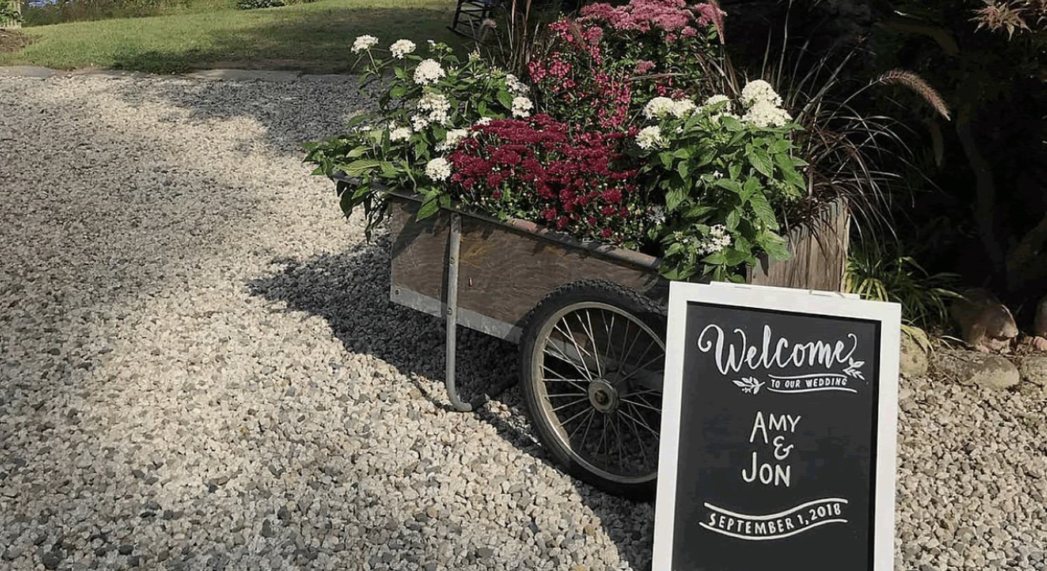 Antique wheelbarrow filled with flowers on gravel with a Welcome to our Wedding sign