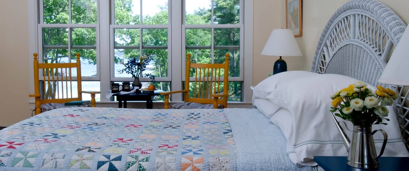 Bright and airy bedroom with bed made up in a colorful quilt with a wicker headboard and two wooden chairs in a seating area.
