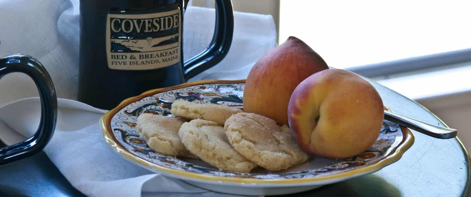 Two ripe peaches and sugar cookies on a plate next to a green coffee mug with the Coveside Bed & Breakfast logo.