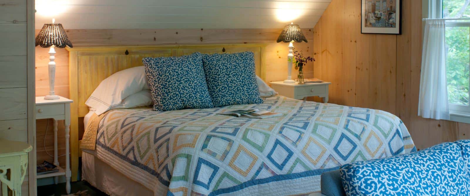 Large bed with a wooden headboard made up with a pretty quilt and blue pillows which match the couch cushions.