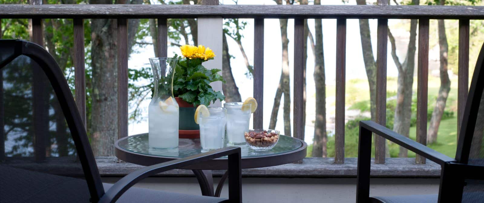 Small glass table holds a carafe of lemonade and two glasses with a bowl of nuts, next to two chairs on a patio.