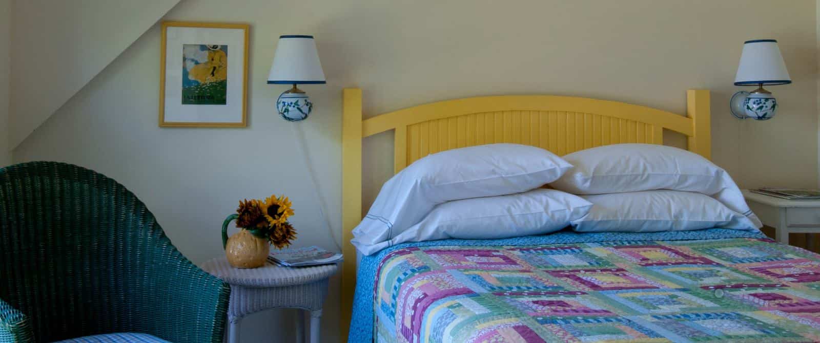 Pretty pastel quilt on a bed with a yellow wooden headboard in bedroom with a green wicker armchair and two china wall sconces.