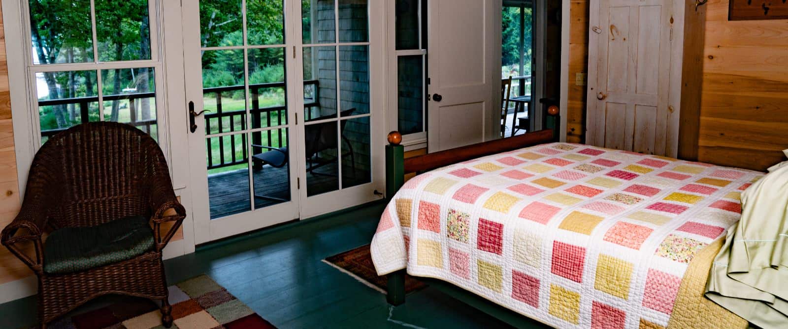 Bedroom with patio doors, green wooden floor, wicker chair and a bed made up in a white, yellow and pink quilt.