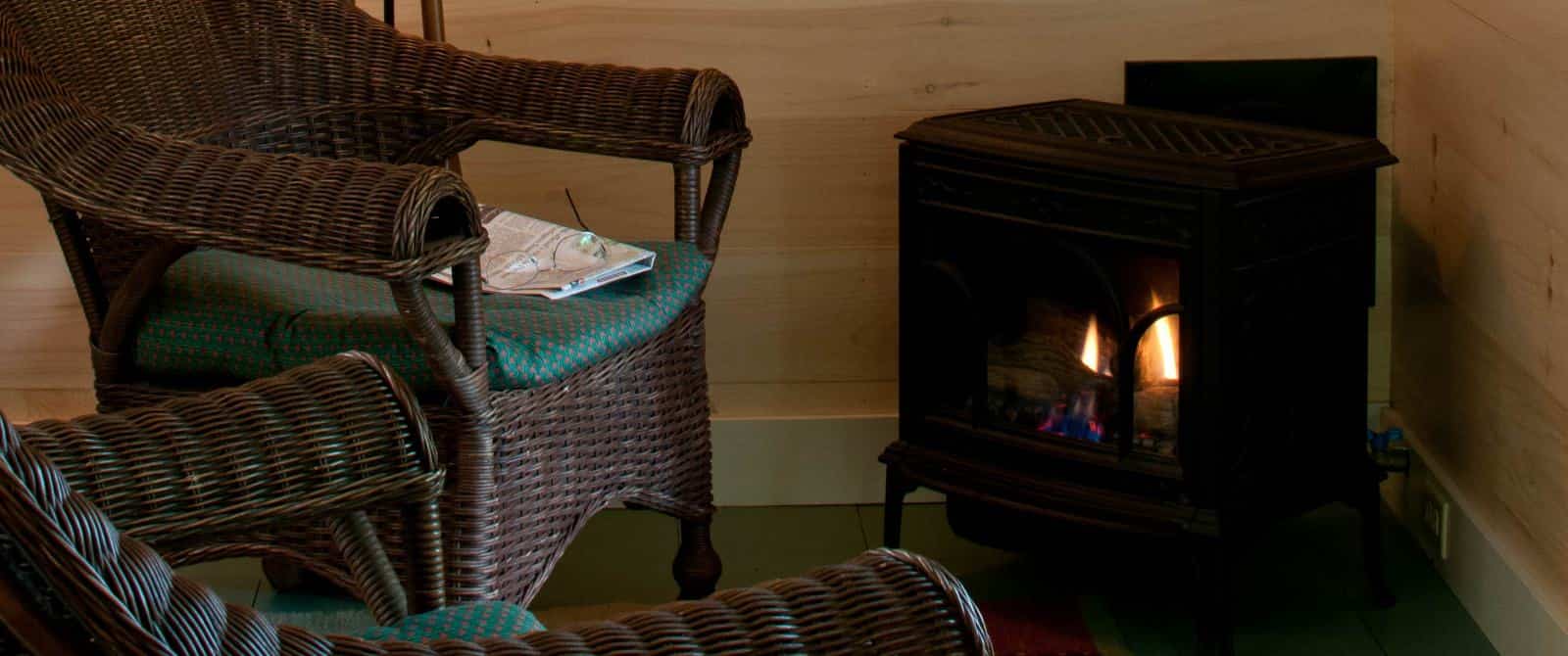 Two wicker armchairs with green padded seats surround a glowing black-iron wood stove.