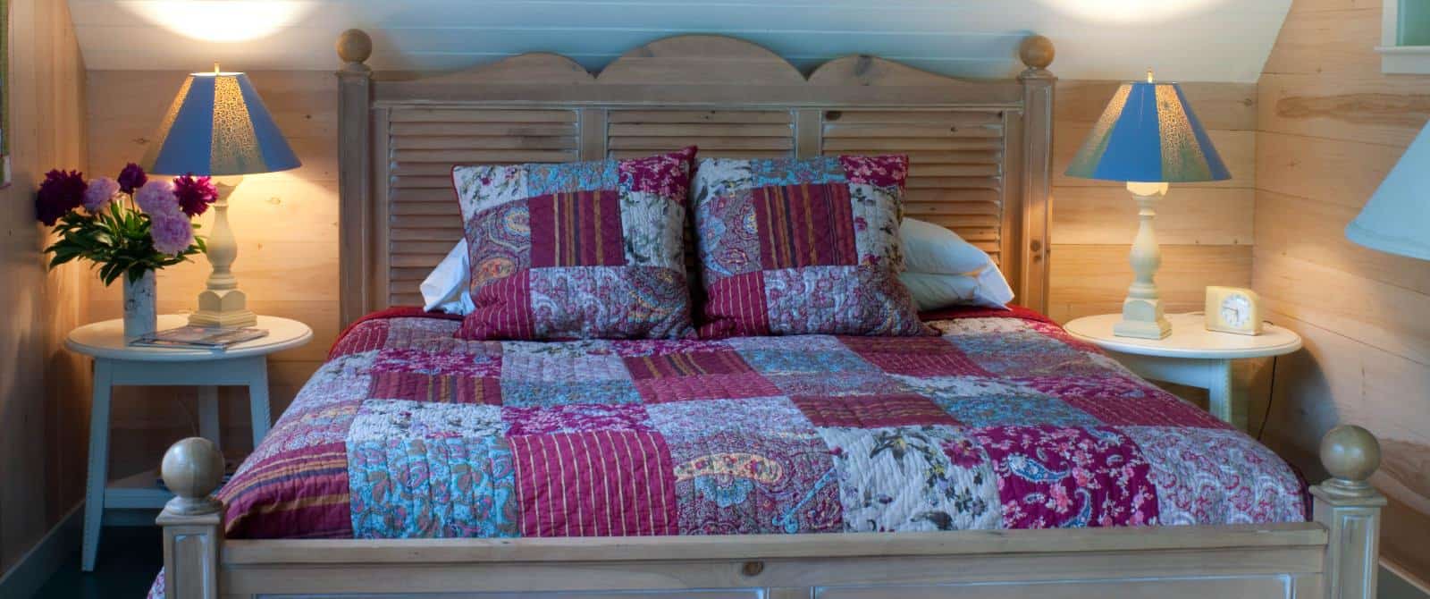 Pretty wooden bed made up with a pink quilt next two two white wooden side table with blue-shaded lamps.