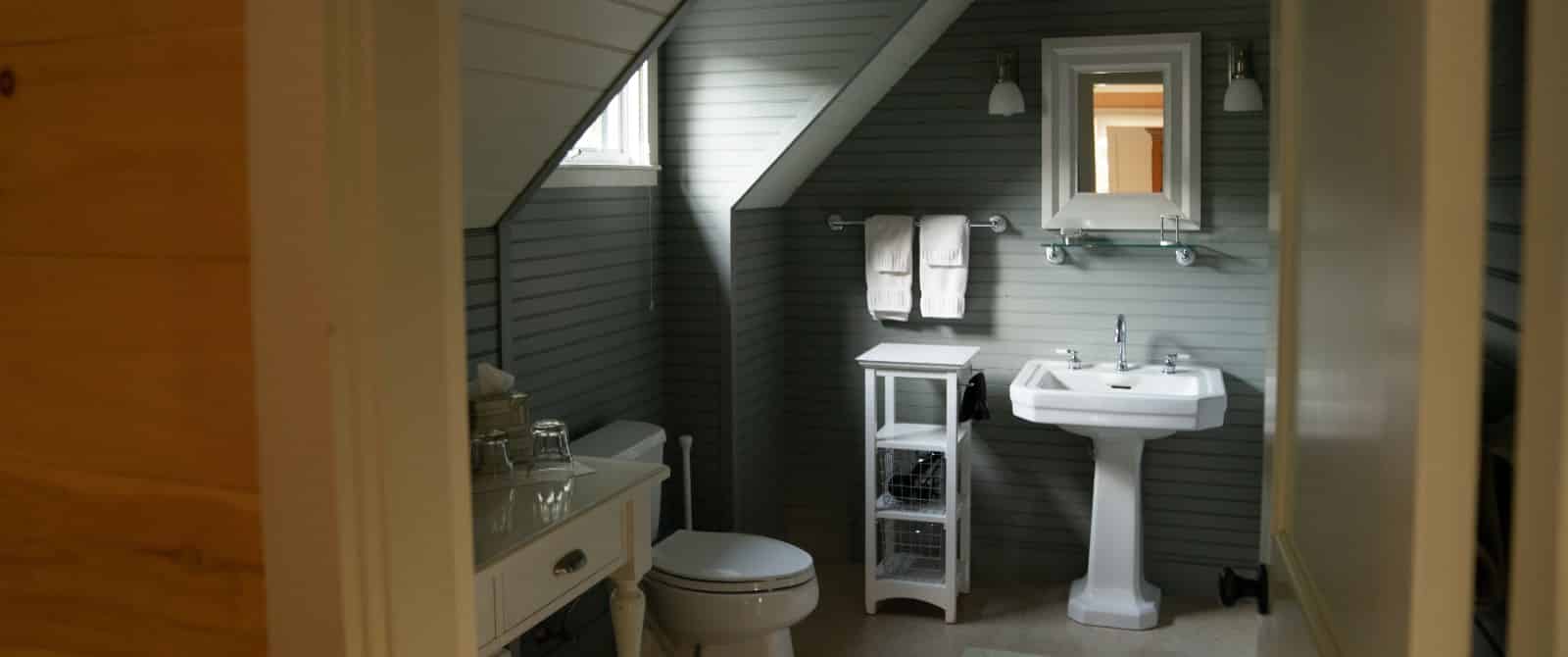 Charming bathroom with sloped ceiling, grey-painted walls, white vanity and two wooden tables.