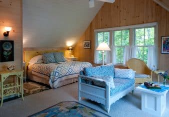 Charming bedroom with sloped ceiling, paneled walls, large bed with a quilt and wooden couch with a small table.
