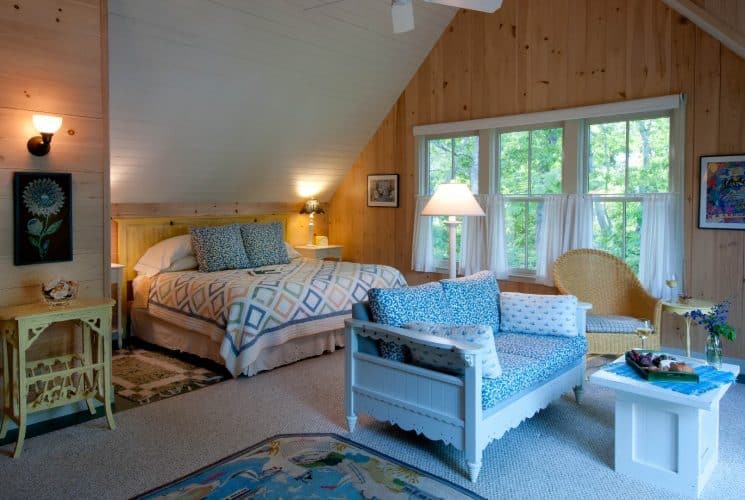 Charming bedroom with sloped ceiling, paneled walls, large bed with a quilt and wooden couch with a small table.