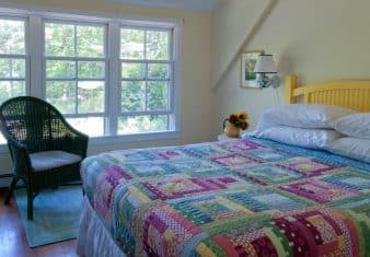 Bright and airy bedroom with three large windows, sloped ceiling and yellow bed made up in a colorful quilt next to a wicker armchair with a padded seat.