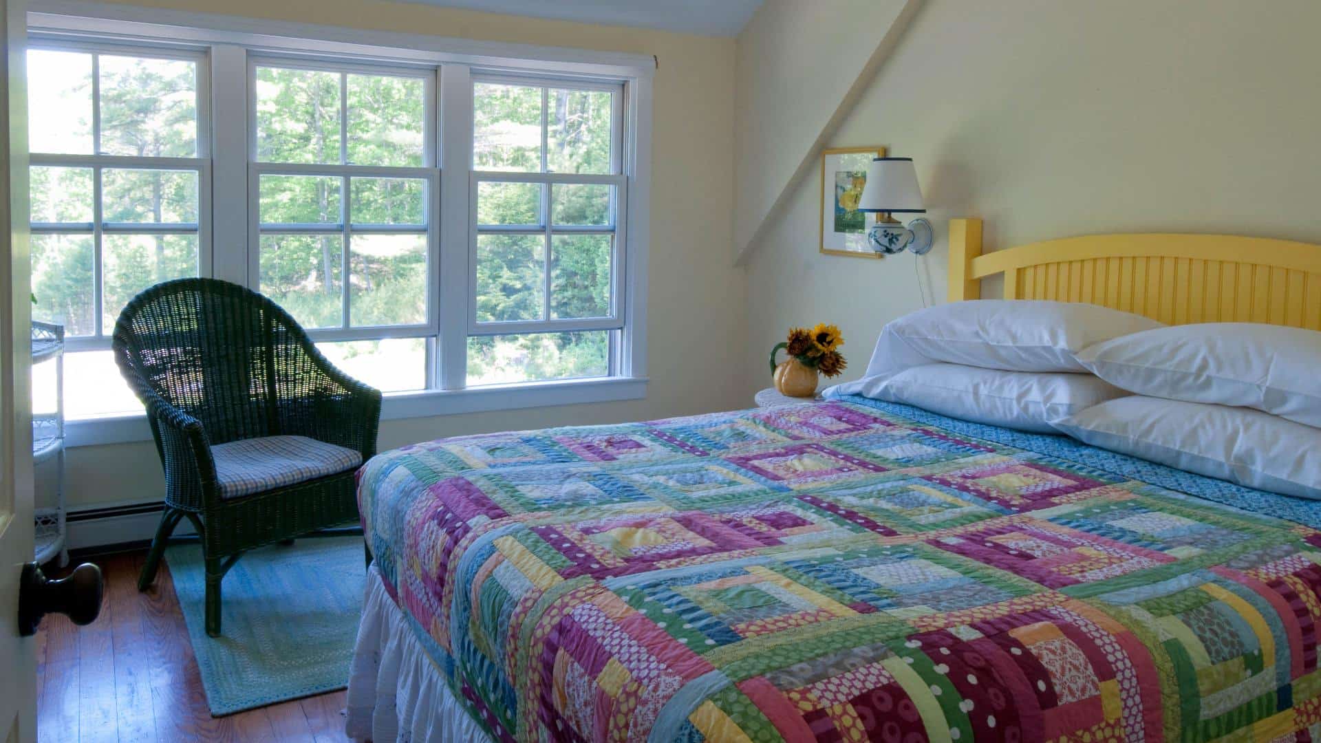 Bright and airy bedroom with three large windows, sloped ceiling and yellow bed made up in a colorful quilt next to a wicker armchair with a padded seat.