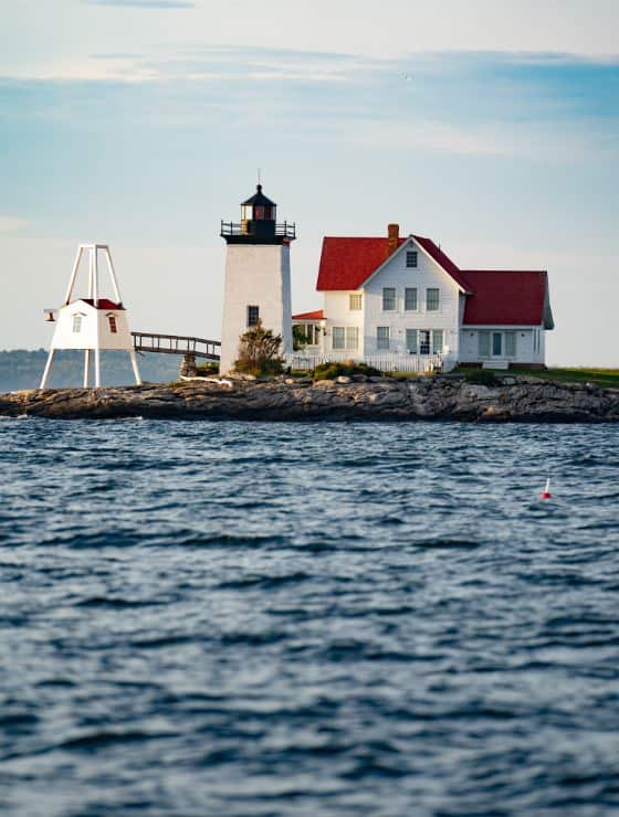Lighthouse and red-roofed keeper's house on stone shoreline in midst of rippling blue water.