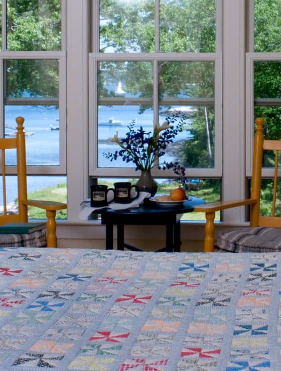 Looking out at the cove from inside a bedroom with large windows, two wooden chairs and bed made up in colorful quilt.