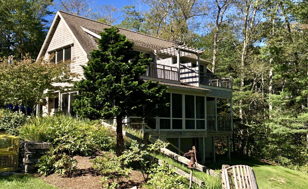 Cottage with tan shake siding and a brown metal roof set among trees, flowering bushes and a low stone wall.